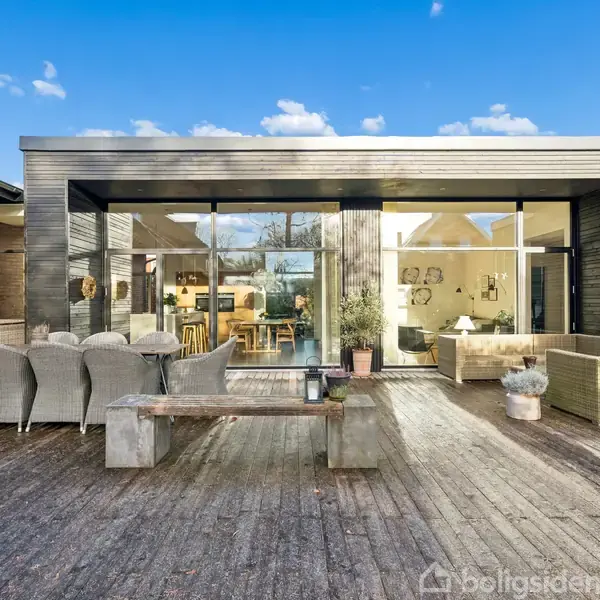 Træterrasse med udendørs spisebord og stole, samt bænke foran store glaspartier. Indenfor ses moderne indretning i et hus med træ- og murstensdetaljer under en blå himmel.
