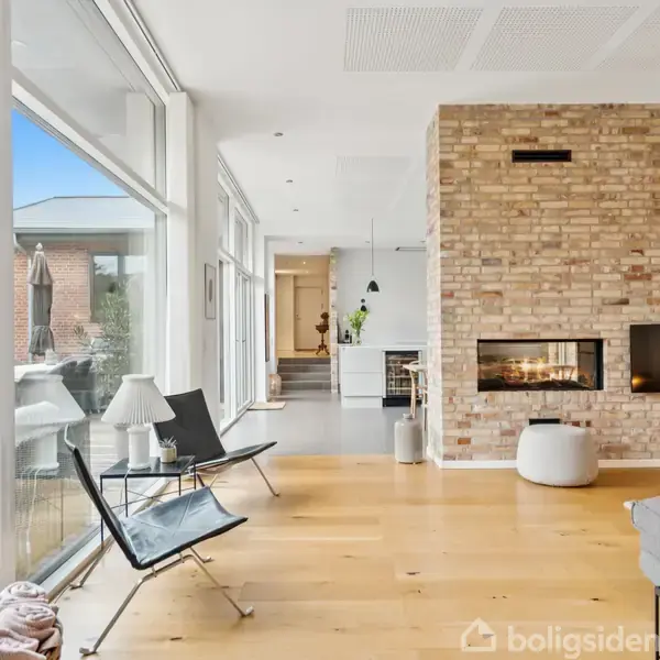 Stue med moderne design; en åben pejs i en murstensvæg, trægulv og store vinduer, der fører ud til en terrasse. To stole og et tæppe er placeret ved vinduet.