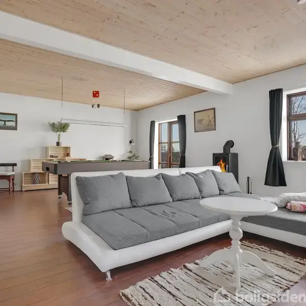 Grå sofa med hvid base står i lys stue med træloft og gulv. Foran sofaen er et rundt bord og et tæppe. Bagerst ses et billardbord og vinduer med sorte gardiner.