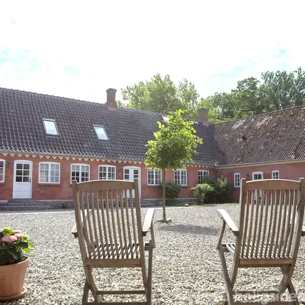 To træstole står på en gruset gårdsplads foran en lyserød bygning med skråt tag og flere vinduer. En ensom træ vokser midt i gårdspladsen.