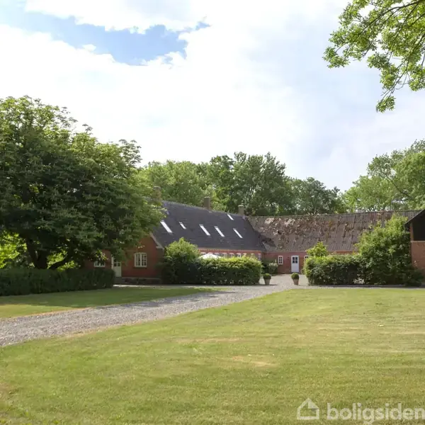 En murstensgård med hvide vinduer ligger stille omgivet af grønne træer og en velplejet græsplæne. En grussti fører op til gården, der ender i en gårdsplads.