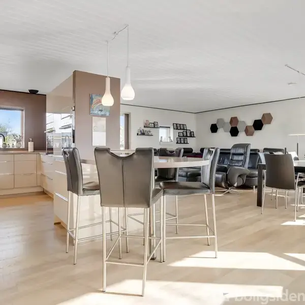 Moderne køkken og opholdsområde med åbent layout. Køkkenø med høje stole til venstre, spisebord med stole til højre. Store vinduer slipper lys ind, og udendørs planter ses udenfor.