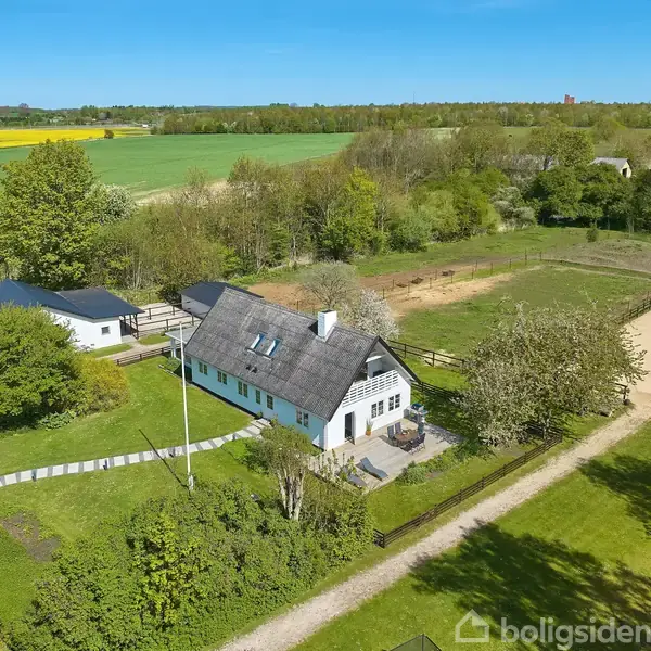 Huset har en stråtækt tag og hvide vægge, beliggende i en grøn landlig have med mark og træer omkring. Tæt på gårdsplads og hestefold.