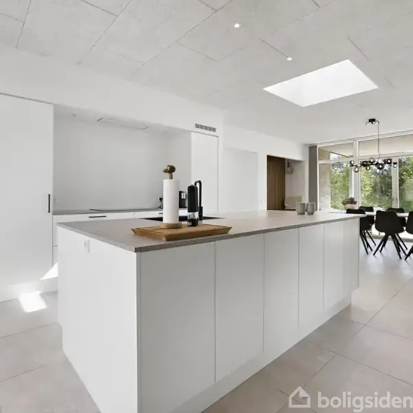 Køkkenø med hvidt bord står centralt. Ovenlysvindue over bordet. I baggrunden ses spisebord og stole ved store vinduer mod en grøn udsigt.