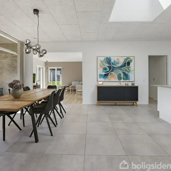 Et rummeligt moderne køkken og spisestue med et stort træbord og stole. Store vinduer åbner mod en terrasse med udsigt over en grøn mark. Kunst hænger på væggen.