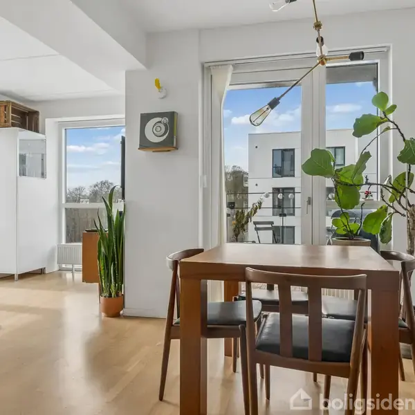 Et spisebord med stole er placeret i et lyst rum med store vinduer. En plante står ved siden af bordet, og der er en guitar og reoler i baggrunden.