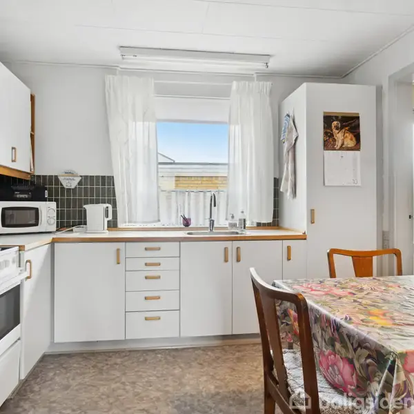 Et lyst køkken indeholder hvide skabe, ovn, mikroovn og elkedel. Et bord med blomsterdug står centralt. Vinduet har udsigt til blå himmel. En kalender hænger ved siden af døren.