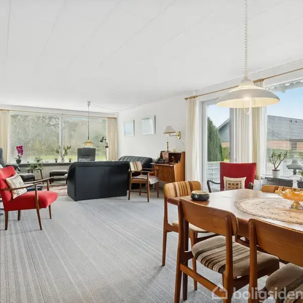 Stue med spisebord og stole i forgrunden, sofagruppe i baggrunden, store vinduer trukket for med lyse gardiner, lys tæppebelægning og reol langs væggen.