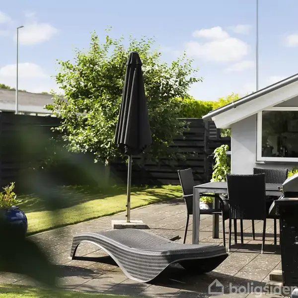 Solstol står på flisebelagt terrasse med et havebord og stole. En sort parasol er slået op. Omgivende have med græsplæne og hæk omkranser området.
