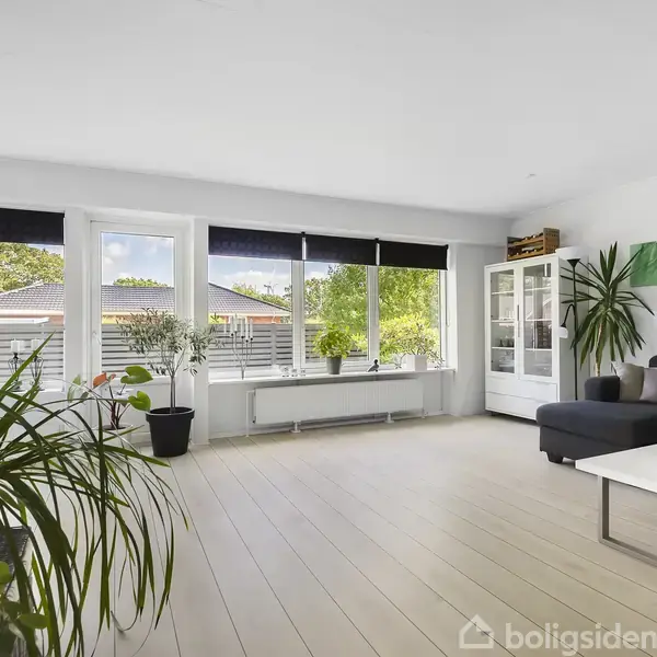Stue med sofa og sofabord i hjørnet, omgivet af grønne planter og kunst på væggen. Store vinduer giver udsigt til en have. Naturligt lys fylder rummet.