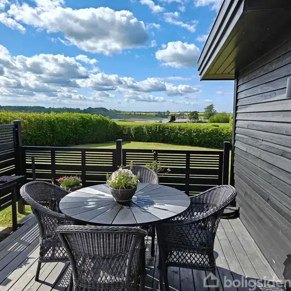 En rund, sort terrassebord med flettede stole står på en træterrasse. Skrånende grønne marker og buske omgiver området under en himmel med spredte skyer.