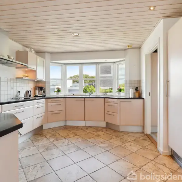 Et moderne køkken med hvide skabe og sorte bordplader. Køkkenapparaterne er indbygget. Stort vindue giver udsigt til en grøn have. Flisegulv og hvidt loft med indbygget belysning.