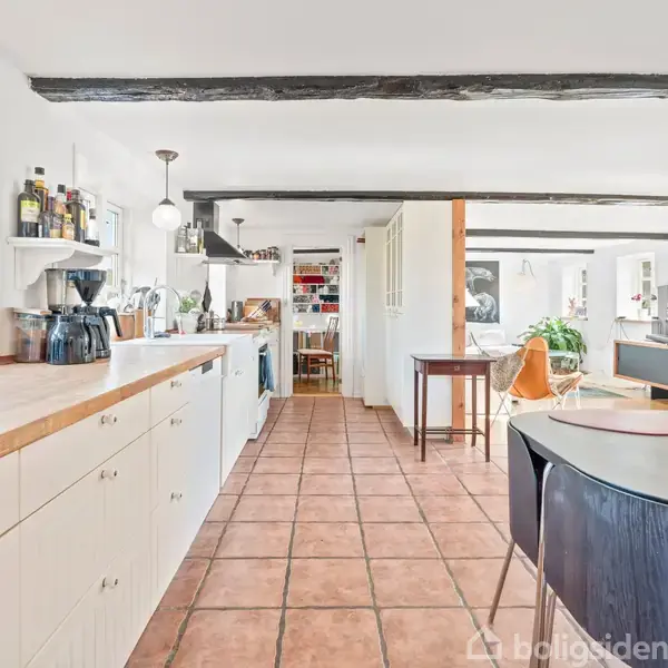 Køkken med hvide skabe og træbordplade. Køkkenet er ryddeligt, med forskellig køkkenudstyr og flasker på bordet. Rummet har lyse fliser og synlige træbjælker i loftet.