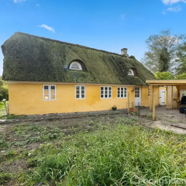 Gult, stråtækt hus med flere små vinduer, står i en have med græs, træer i baggrunden og en overdækket terrasse på højre side. Udsigt under en klar blå himmel.