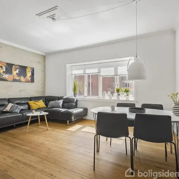 En sort lædersofa står i en stue ved siden af et rundt bord med stole. Rummet har et stort vindue, træparketgulv og moderne kunst på væggen.