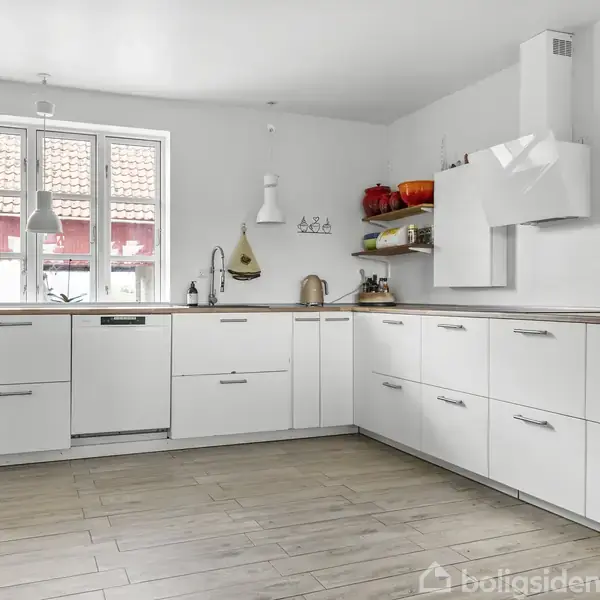 Hvidt køkken med moderne skabe og skuffer, placeret omkring en hjørneopsætning. Træbordplader og hylder med rød gryde og krydderiplanter. Rummet er lyst med vinduer mod en solrig gård.