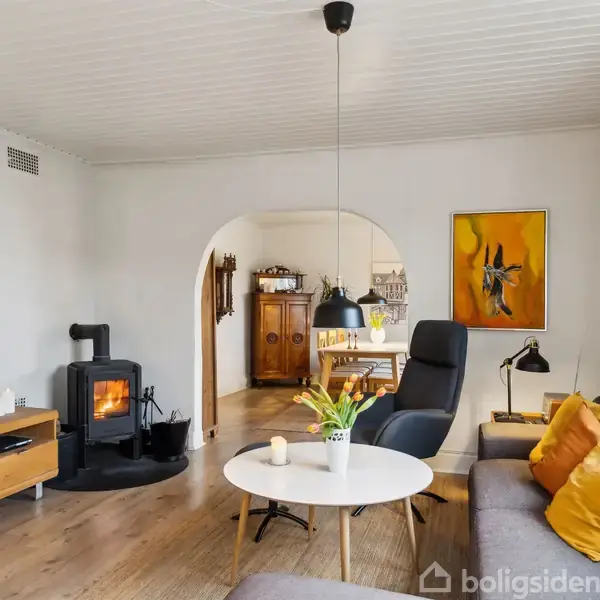 En stue med en sofa og lænestol har puder i gule nuancer. Et rundt sofabord har en vase med blomster. En brændeovn brænder ved siden af et TV på et træmøbel. En lysbue fører til spisestuen med et skab. En farverig maleri hænger på væggen.