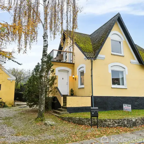 Et gult hus med spidst tag står forrest. Huset har en balkon og trapper ved indgangen. Til venstre ses en gul garagebygning. Et skilt med teksten "Til salg RealMæglerne Espergarde" står foran huset. Træer omgiver omgivelserne.