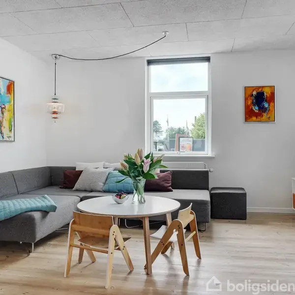 Gråt hjørnesofa arrangement med sofabord og blomster i vase, i en stue med lyse trægulve og farverige kunstværker på væggene. Store vinduer lader naturligt lys strømme ind.