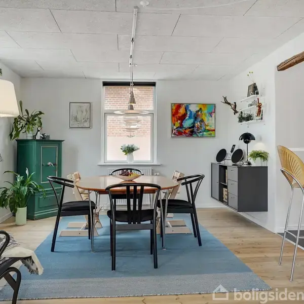 Spisebord med seks stole står på et blåt tæppe i en lys spisestue. Omgivet af grønne planter, moderne kunst på væggen, en grøn skænk og en disk med barstole.