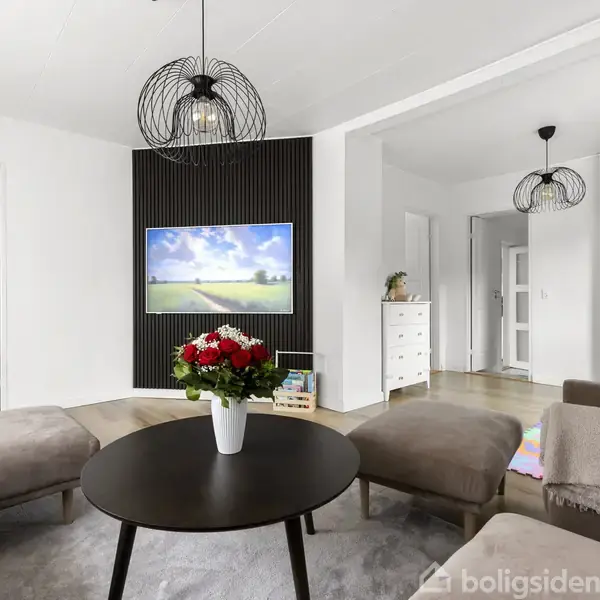 Stue med grå sofaer omkring et rundt bord med blomster. Et fjernsyn viser en naturudsigt. Moderne lamper hænger ned fra loftet, og der er sorte paneler på væggene.