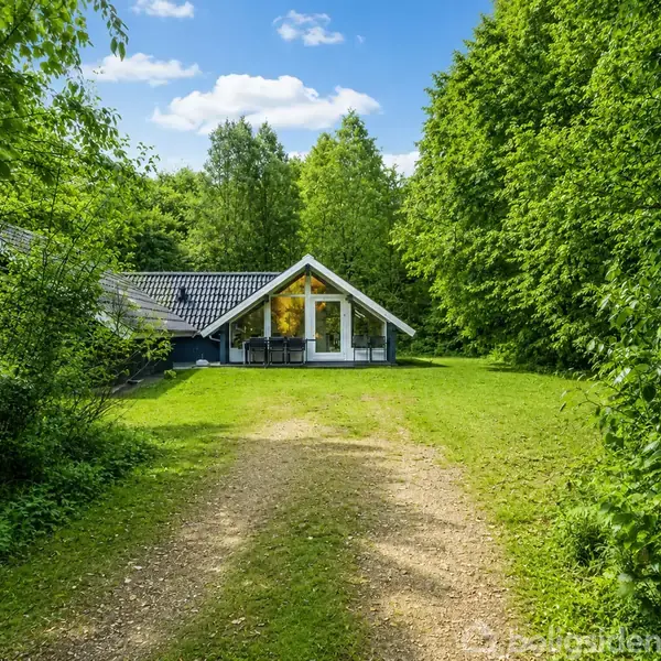 Hus med spidstag står på en grusvej, omringet af tæt grøn skov. Store vinduer reflekterer himlen, og en velholdt græsplæne strækker sig foran.