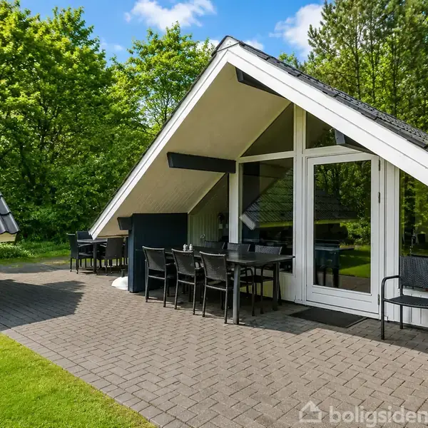 Et sommerhus med trekantet tag står på en stenbelagt terrasse. Der er et udendørs spisebord med stole. I baggrunden er der frodig skov. Nummer 114 ses på huset.