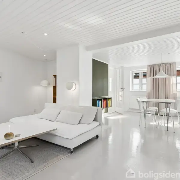 En hvid sofa står i en lys, minimalistisk stue med et lavt sofabord foran. Spisestuen i baggrunden har et rundt bord og stole nær et vindue med gardiner.