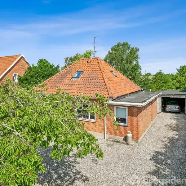 Et rødt murstenshus med orange tegltag står stille ved siden af en carport i en grusbelagt indkørsel. Træer og grønne buske omkranser huset under en klar blå himmel.