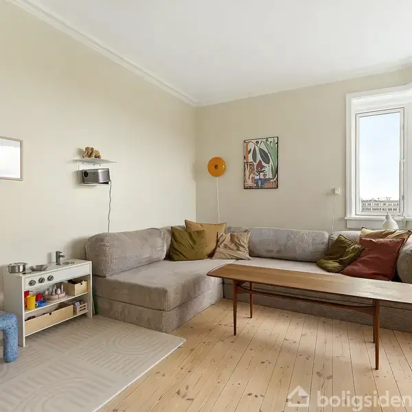 En grå hjørnesofa med mange puder står i en lys stue med lyse vægge. Et træbord står foran sofaen. Der er billeder og en hylde på væggene.