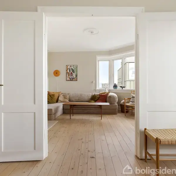 En stue med en åben dobbelt dør viser en sofa med puder i forskellige farver. Rummet har lyse trægulve, vægkunst og en stor vindueskarm med udsigt.