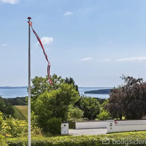 Flagstang med flagrende vimpel i forgrunden, omgivet af grønne buske og træer. I baggrunden ses et hvidt hus og en blå fjord under en klar himmel.