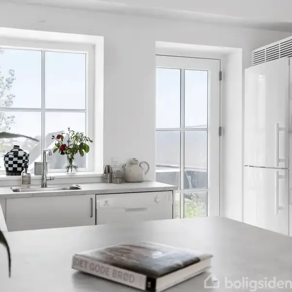 Et moderne køkken med hvide skabe og en integreret ovn. En bog med teksten "DET GODE BRØD" ligger på bordet. Lyse vinduer giver et luftigt indtryk.