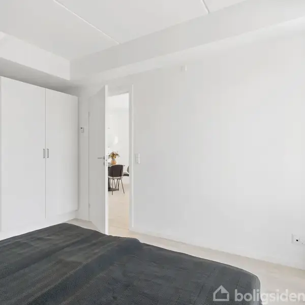 Seng står i et lyst soveværelse med hvidt klædeskab ved siden af. Døren fører til en spisestue, og en plante står i hjørnet.