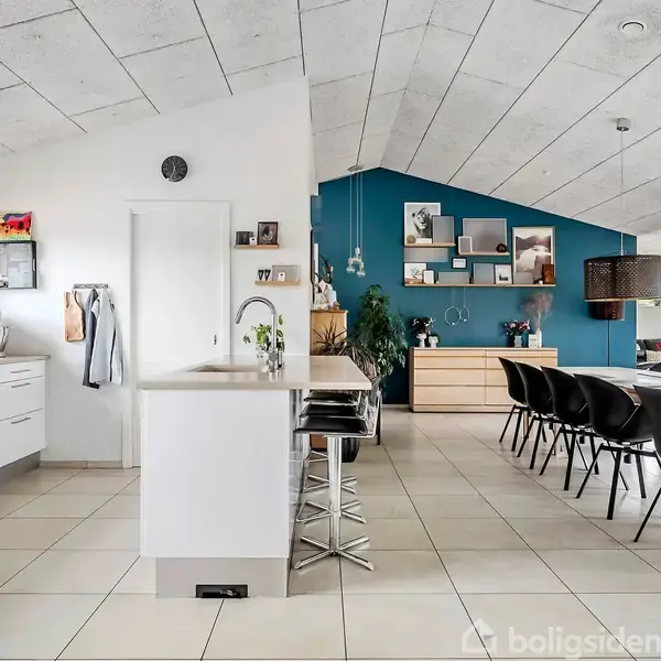Et moderne køkken med hvide skabe og en køkkenø. To barstole står ved øen. I baggrunden ses en spisestue med sort spisebord og blå væg.