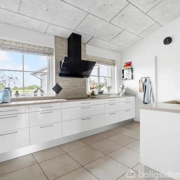 Et lyst, moderne køkken med hvide skabe, en sort emhætte, og et lyst klinkegulv. Store vinduer lukker dagslys ind. Emhætten hænger centralt, omgivet af en minimalistisk indretning.