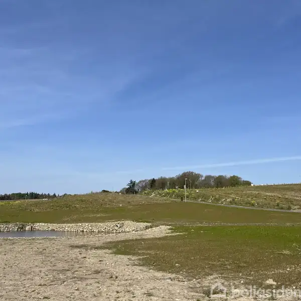 Åben mark med en lille vandpyt, græsklædt areal og en grusvej, omgivet af let kuperet terræn og træer i baggrunden, under en klar blå himmel.