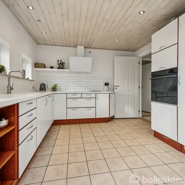 Køkken med hvide skabe og skuffer, udfører funktion som madlavningsområde, omgivet af fliser på væggene og lys trægulv. Hylder viser kopper og små planter.