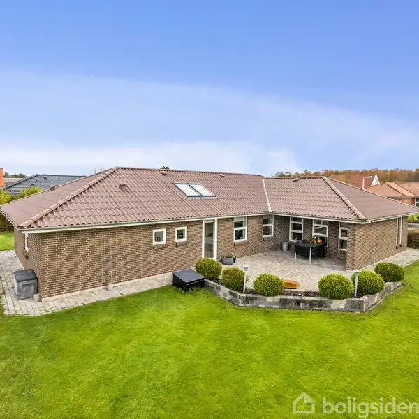 Et moderne etplanshus ligger på en velplejet græsplæne. Huset har murstensfacade og rødt tegltag. En terrasse med havemøbler er synlig, omgivet af velplejede buske.
