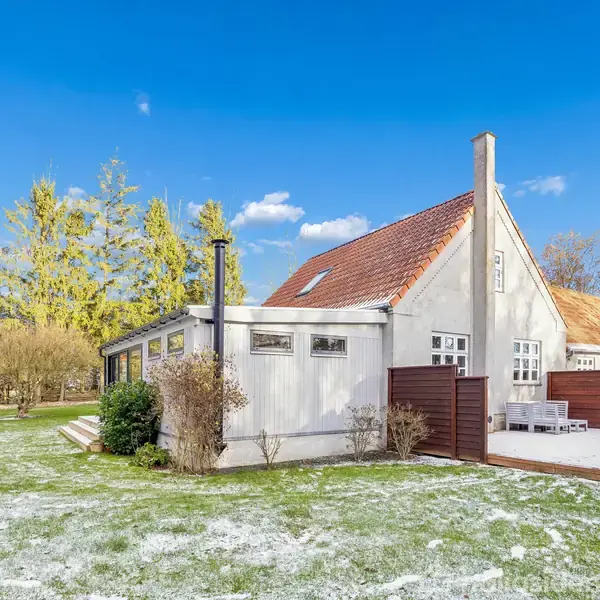 Et hvidt hus med rødt tegltag står i en have med spredt sne. Ved siden af huset er der en terrasse med havehegn, og der er træer i baggrunden under en blå himmel.