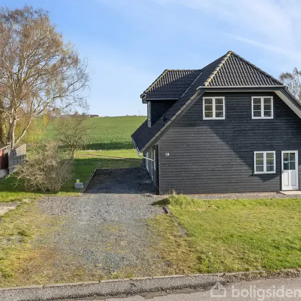 Sort træhus står stille på grusvej, omgivet af grønne plæner og buske, med marker i baggrunden under en blå himmel. To affaldsbeholdere står til venstre.
