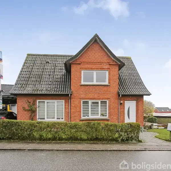 Rødt murstenshus står stille bag en lav hæk langs vejen, med en carport til venstre. Omkring huset ses naboer og en grøn have. φωτοgrafierne på lygtepælen er valgplakater.