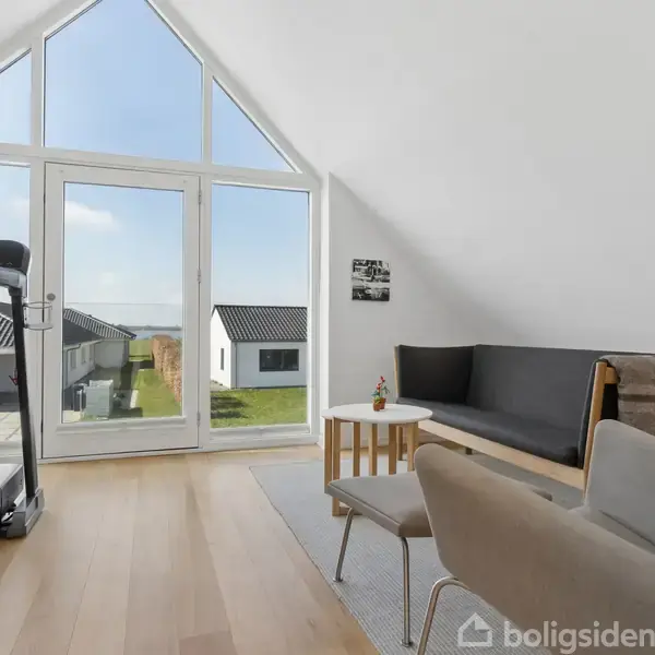 Lys stue med en træningsmaskine og moderne møbler foran en stor vinduesvæg. Udsigt til et kvarter med lave bygninger. En plante findes på et lille bord.