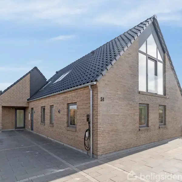 En moderne, lys murstensvilla med skråt, mørkt tag. Huset har store vinduer og en indkørsel med fliser. Der er tekst "51" på husvæggen. Omkring er der hæk og flisebelagt terrasse.
