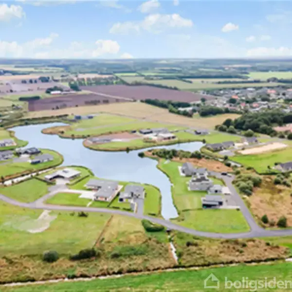 Boligområde med spredte huse omgivet af vejforløb og en snoet sø, beliggende i et landområde med marker i baggrunden under en blå himmel med skyer.