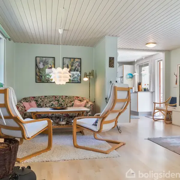 Stue med træstole og blomstrede sofa omkring et sofabord. Brændeovn til venstre, lys hængelampe over bordet. Malerier på væggene, lys grøn vægmaling.