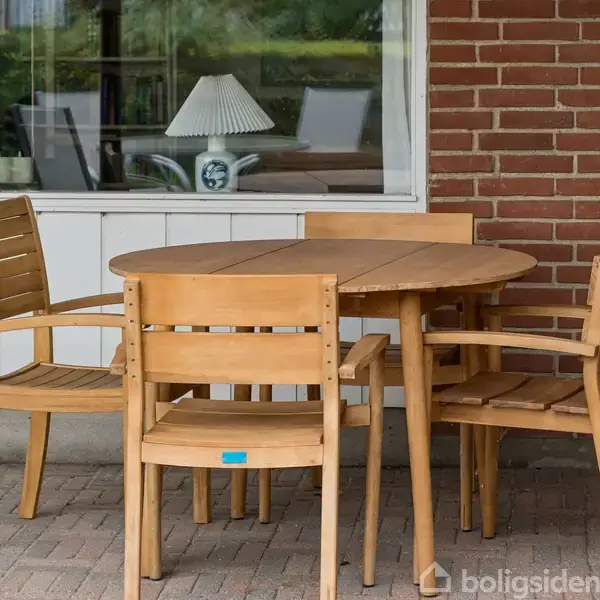 Et rundt træbord med fire tilhørende stole står på en flisebelagt terrasse foran et hus med røde mursten og et vindue. Indendørs ses en lampe på et bord.