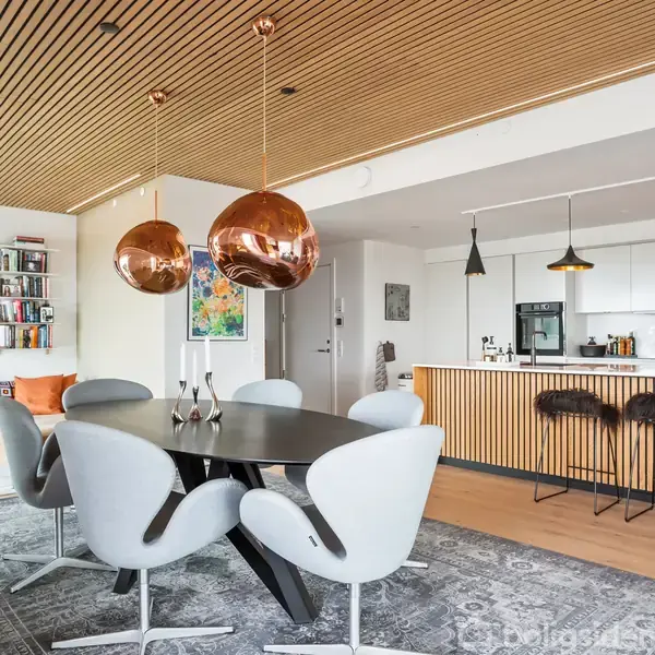 Et moderne spiseområde med seks hvide stole omkring et sort bord. I køkkenet er der en hvid indretning med trædetaljer og to kobberlamper hængende over bordet.
