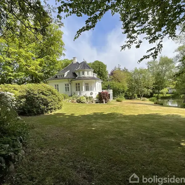 Et hvidt, charmerende hus står omgivet af frodig have med græsplæne og buske. Træer skaber en idyllisk atmosfære, mens en lille sø ses i baggrunden under en blå himmel.
