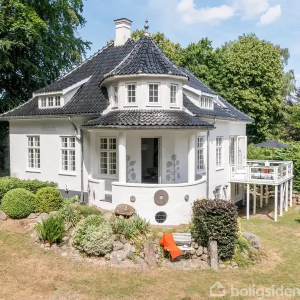 Hvid villa med mørkt tegltag står i en frodig have. Huset har en rund karnap og flere vinduer. Omkring er der buske, træer og en græsplæne.